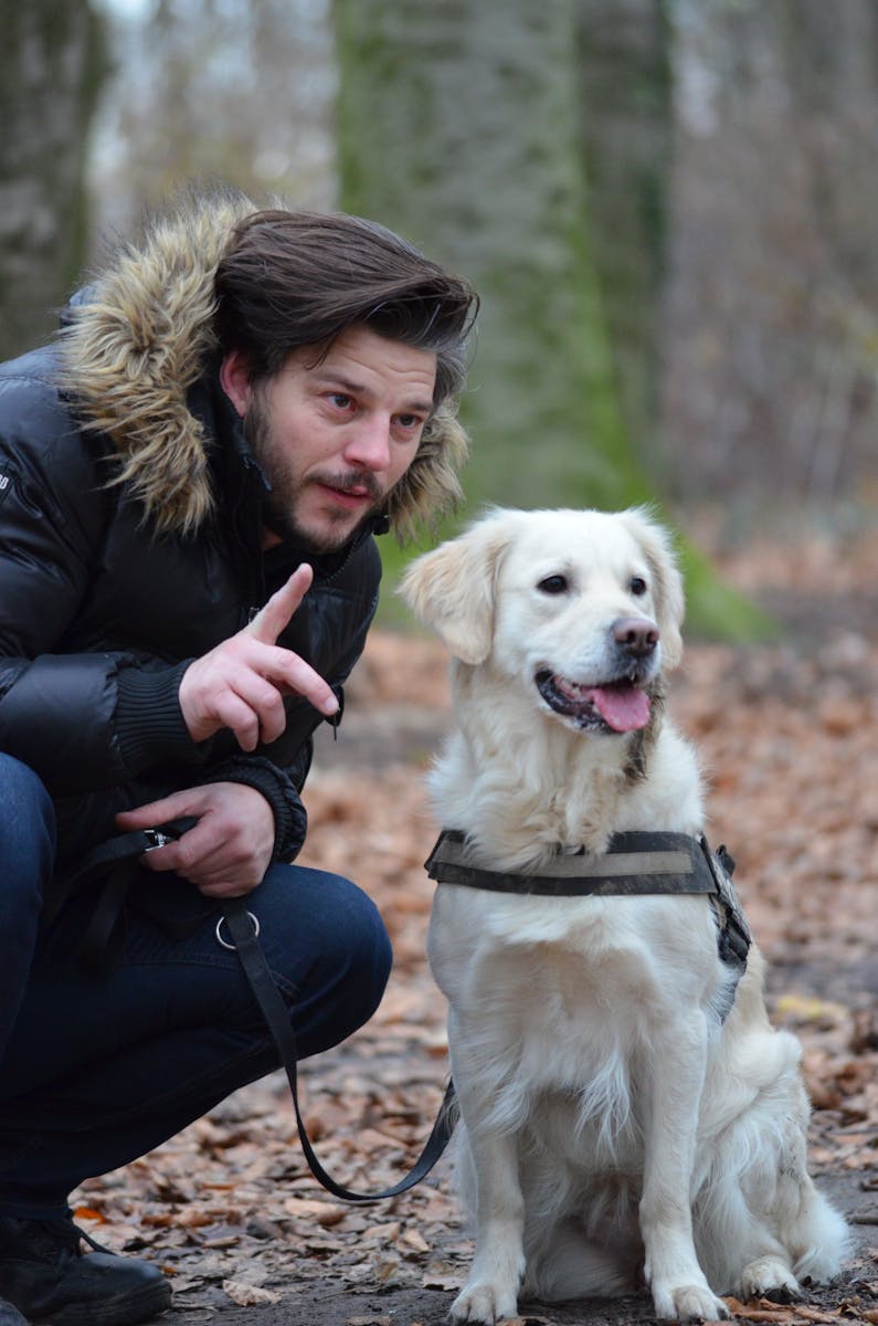 Man Wearing Black and Brown Fur Hoodie Jacket and Blue Pants Holding Dog Leash Beside White Short Coat Dog